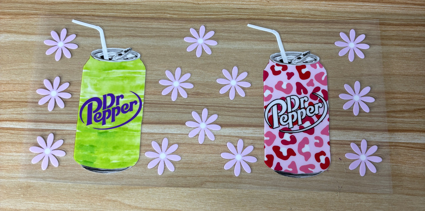 two cans of Dr Pepper soda, one green and one pink, with straws sticking out of them, surrounded by pink flowers on a wooden surface.