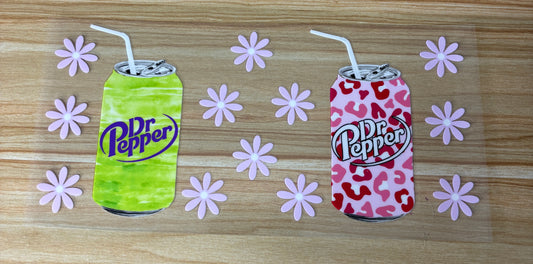 two cans of Dr Pepper soda, one green and one pink, with straws sticking out of them, surrounded by pink flowers on a wooden surface.